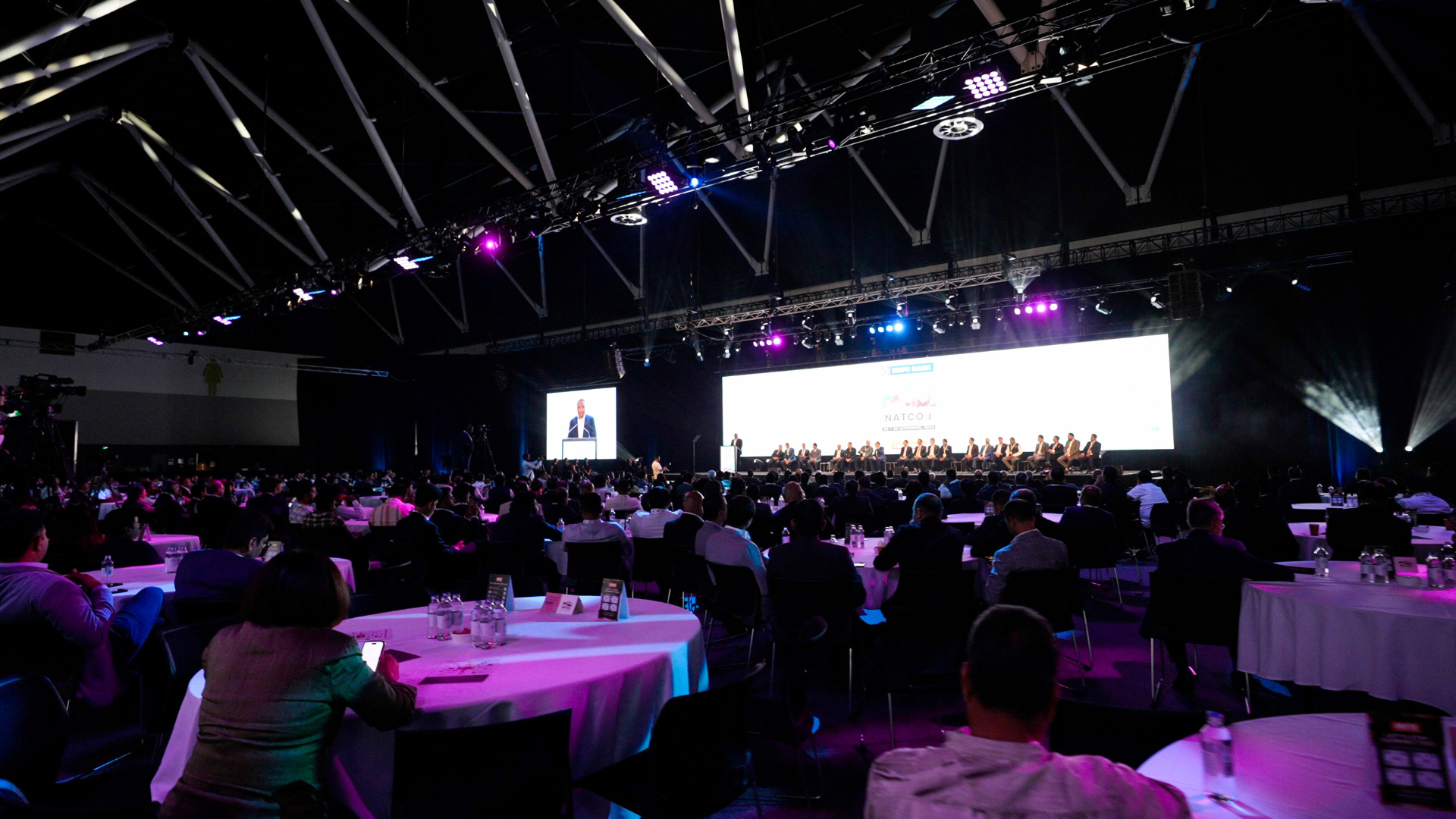 View from back of room full of seated delegates and speaker panel 
