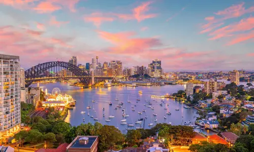 Dusk view of Sydney Harbour with pink and blue skies over the Harbour Bridge and Luna Park. City lights are golden.