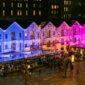 Crowds enjoying outdoor dining at Campbell's Stores at night, illuminated with vibrant blue and pink light projections