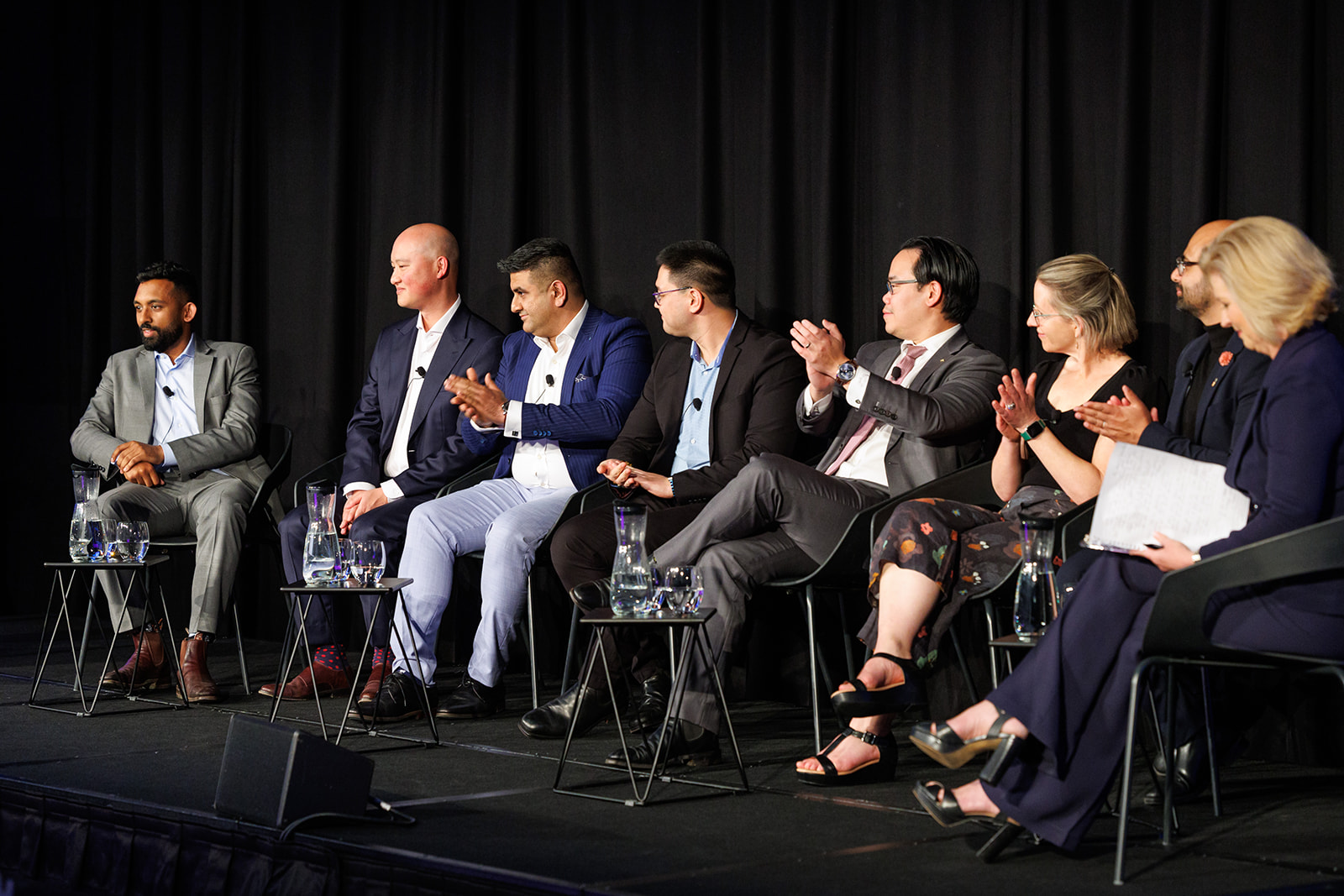 A Panel Of Seven People Seated On A Stage Participating In A Formal Discussion