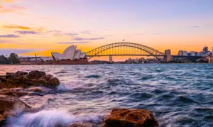 Waves over rocks with the Harbour Bridge and Opera House in the background at sunset