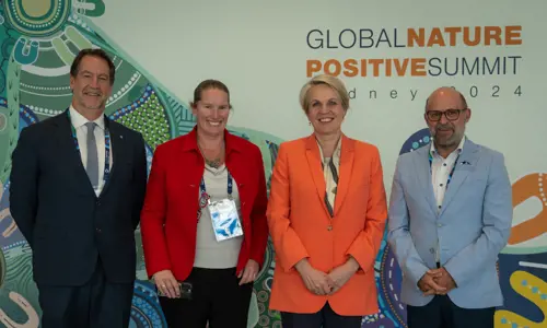 Four Conference Attendees Standing In Front Of Large Entrance Board With The Title Name Of Conference Global Nature Positive Summit Logo