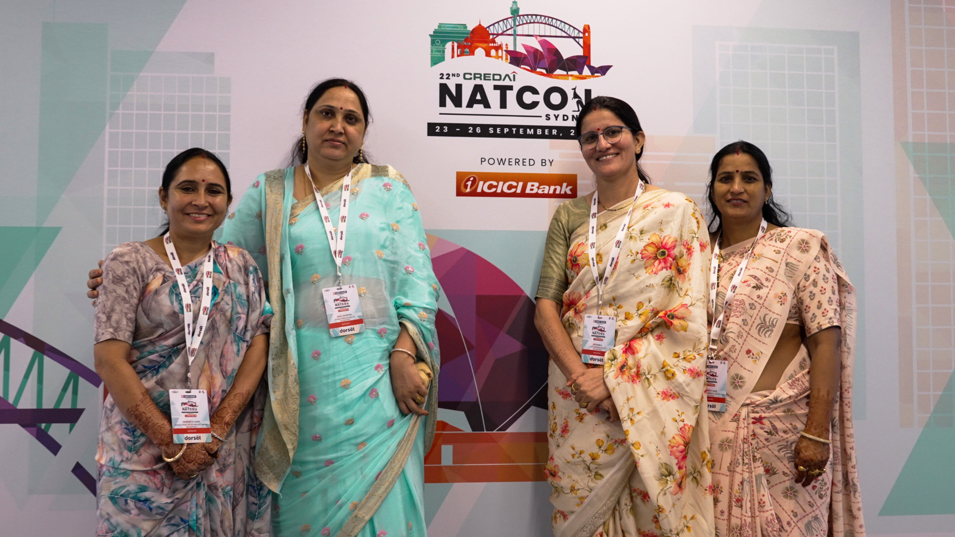 Group of four women delegates in traditional sari outfits, standing in front of the Credai Natcon signage board at entrance